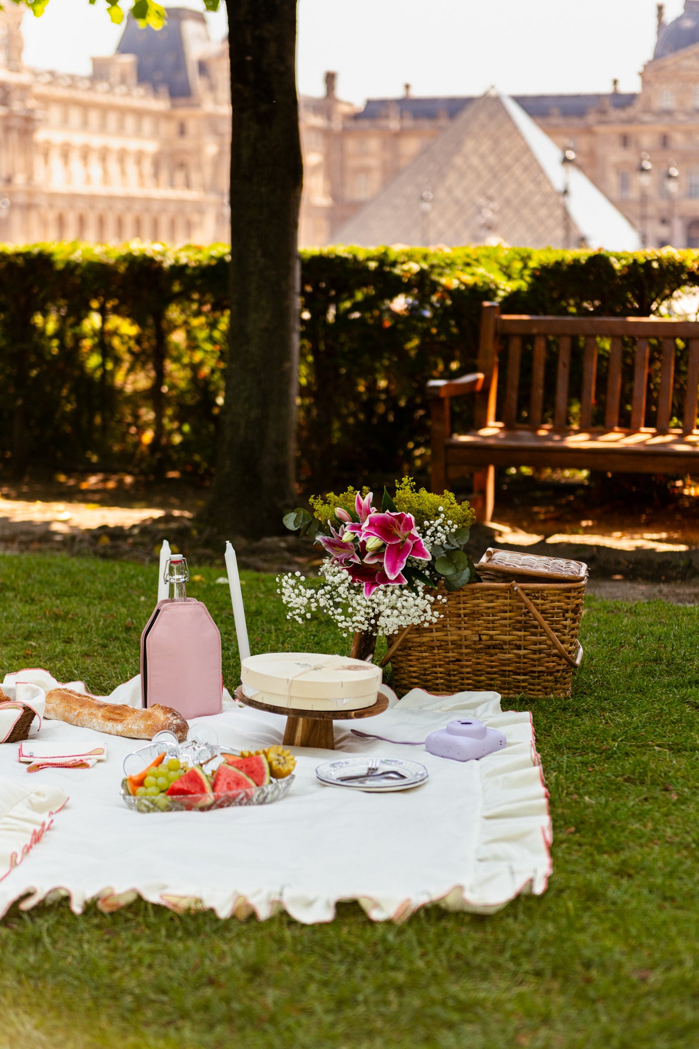 A beautifully arranged picnic setup in Paris with a checkered blanket, a wicker basket, fresh baguette, fruits, and wine, set against the backdrop of a Parisian park.